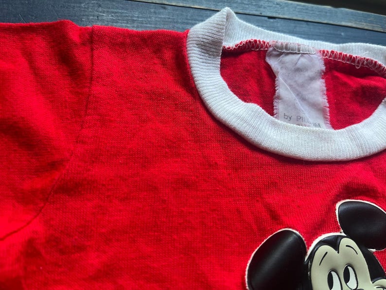 K&ouml;nnte beinhalten: Ein rotes T-Shirt mit wei&szlig;em Kragen und einer Mickey Mouse-Grafik. Das Shirt hat ein Etikett mit der Aufschrift "by P...". Das Shirt ist aus einem weichen Material gefertigt.