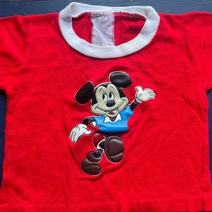 K&ouml;nnte beinhalten: Ein rotes T-Shirt mit wei&szlig;em Kragen, das eine gro&szlig;e Grafik von Mickey Mouse zeigt. Mickey ist in einer dynamischen Pose dargestellt und tr&auml;gt ein blaues Hemd und braune Schuhe. Das Shirt ist wahrscheinlich f&uuml;r Kinder.