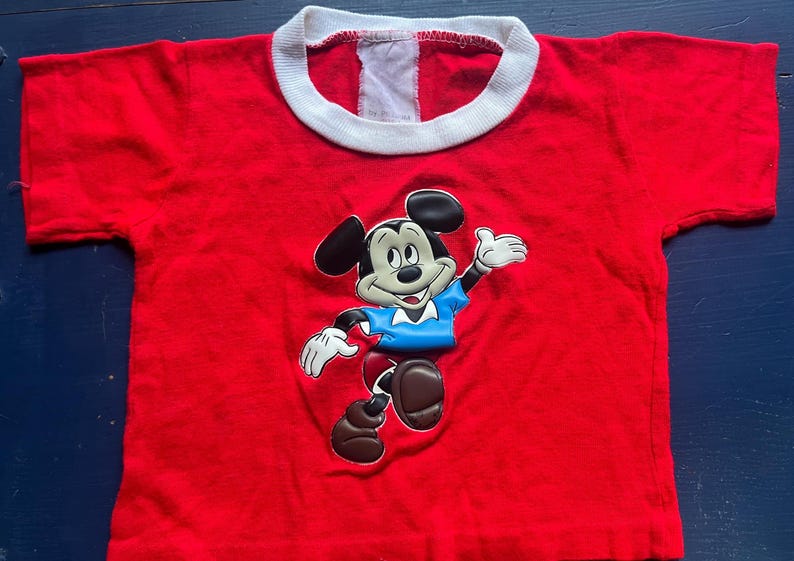 K&ouml;nnte beinhalten: Ein rotes T-Shirt mit wei&szlig;em Kragen, das eine gro&szlig;e Grafik von Mickey Mouse zeigt. Mickey ist in einer dynamischen Pose dargestellt und tr&auml;gt ein blaues Hemd und braune Schuhe. Das Shirt ist wahrscheinlich f&uuml;r Kinder.