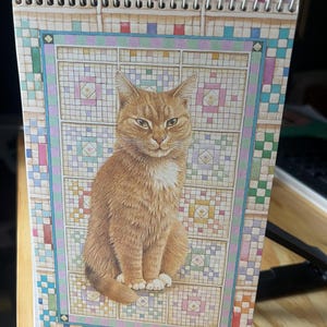 May include: A spiral-bound notebook featuring a detailed illustration of an orange tabby cat with a white chest patch sitting in front of a colorful, grid-patterned quilt design with small square blocks in shades of pink, blue, and yellow.