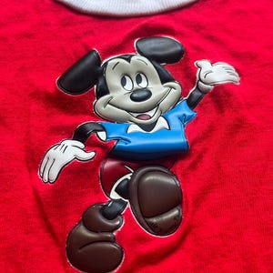 K&ouml;nnte beinhalten: Ein rotes T-Shirt mit einem gro&szlig;en, erhabenen Aufdruck von Mickey Mouse. Mickey ist in seiner klassischen Kleidung dargestellt: schwarze Ohren, blaues Hemd, rote Shorts und braune Schuhe. Das T-Shirt hat einen wei&szlig;en Kragen.