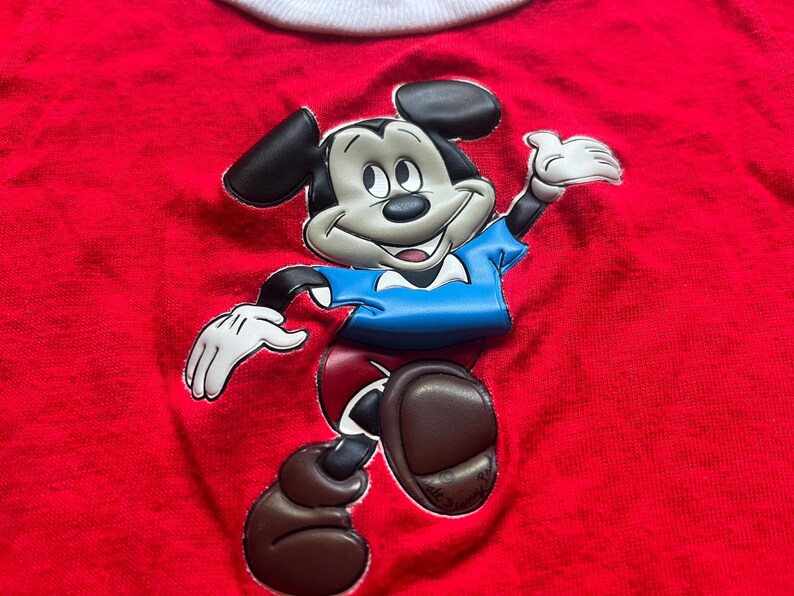 K&ouml;nnte beinhalten: Ein rotes T-Shirt mit einem gro&szlig;en, erhabenen Aufdruck von Mickey Mouse. Mickey ist in seiner klassischen Kleidung dargestellt: schwarze Ohren, blaues Hemd, rote Shorts und braune Schuhe. Das T-Shirt hat einen wei&szlig;en Kragen.