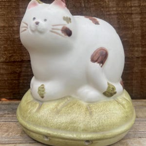 May include: A ceramic cat figurine with a white body and brown spots, sitting atop a light green cushion-shaped base. The cat has pink accents on its ears and a smiling face. The base has small holes.