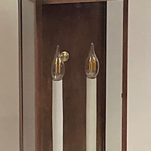May include: A copper-colored outdoor wall sconce with a rectangular glass enclosure. Inside are two white candle-shaped lights with brass-colored bases and a curved top. The sconce is designed for exterior lighting.