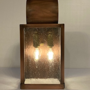 May include: A copper-toned wall sconce with a curved top and a rectangular glass panel. The glass has a textured, bubbled effect and two light bulbs are visible inside.