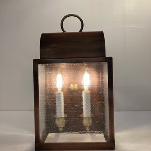 May include: A brown metal outdoor wall lantern with a clear glass pane and two lit bulbs. The lantern has a curved top with a ring handle.