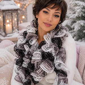 May include: A ruffled scarf featuring a gradient of black, grey, and white tones with an open-knit, lace-like texture. The scarf is worn over a white long-sleeved sweater and is paired with large gold hoop earrings, creating a layered appearance.