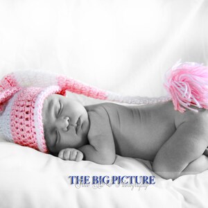 Op de afbeelding: Een slapende pasgeboren baby met een roze en witte gehaakte muts met een lange staart en een roze pompon. De afbeelding is in zwart-wit, met de muts in kleur. De baby ligt op een wit kussen. De tekst "THE BIG PICTURE Fine Art Photography" staat onderaan.
