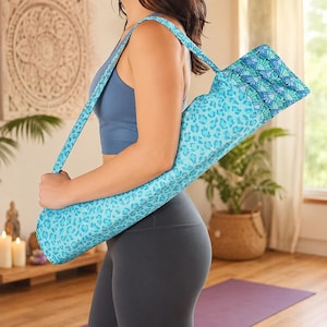 May include: A cylindrical yoga mat bag featuring a light blue and turquoise leopard print pattern on the main body, with a contrasting geometric fan-shaped design in shades of blue and green on the top flap and a matching shoulder strap.