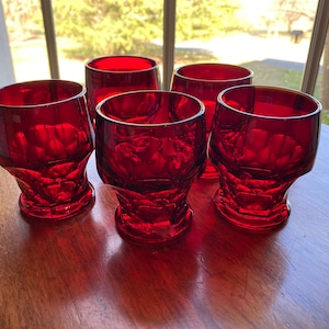 May include: Set of five ruby red drinking glasses. Each glass has a unique, textured design on the lower half and a smooth rim. The glasses are arranged on a wooden surface, with natural light illuminating them from a nearby window.