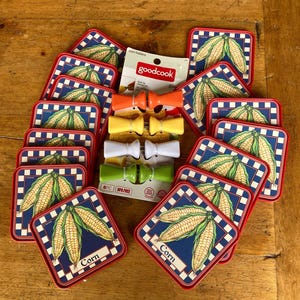 May include: A collection of corn-themed kitchen accessories. Includes square coasters with corn illustrations, red borders, and blue checkered backgrounds. Also, colorful corn holders in orange, yellow, white, and green. The packaging reads "goodcook".