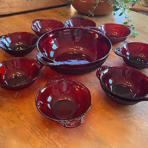 May include: A set of ruby red glass bowls, including a large serving bowl and several smaller bowls with handles. The bowls are arranged on a wooden surface, showcasing their glossy finish and vintage design. The bowls are ideal for serving soups or desserts.