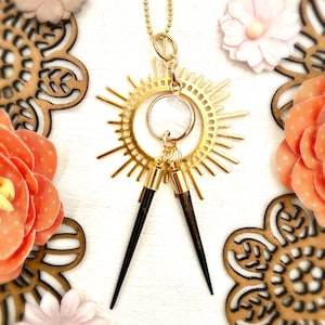 May include: A gold-colored pendant necklace featuring a sunburst design with a circular, iridescent center. Two black, pointed pendants hang below. The necklace is displayed with decorative flowers and wooden cutouts.