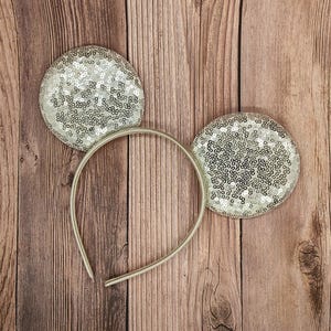 Puede incluir: Diadema con orejas de ratón con lentejuelas plateadas. Las orejas son redondas y están cubiertas de brillantes lentejuelas plateadas. La diadema es de metal fino plateado. Las orejas están unidas a la diadema con una banda plateada. El fondo es una superficie de madera.