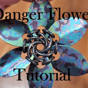 May include: A close-up of a metal flower with a black and blue iridescent finish. The flower is made of multiple petals and has a black metal center. The text "Danger Flower Tutorial" is written in black letters on a brown background.