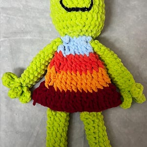 May include: A green crocheted frog wearing a multi-colored dress with a red, orange, yellow, blue, and light blue pattern. The frog has black eyes and a black smile.
