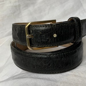 Op de afbeelding: Een zwarte leren riem met een gouden gesp. De riem heeft een getextureerd, gegraveerd patroon.