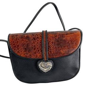 Op de afbeelding: Zwarte en bruine crossbodytas met een klep met textuur van imitatiekrokodil. De tas heeft een zilverkleurige hartvormige sluiting en een zwarte riem. De tas is ontworpen met een gebogen vorm en een enkele riem.