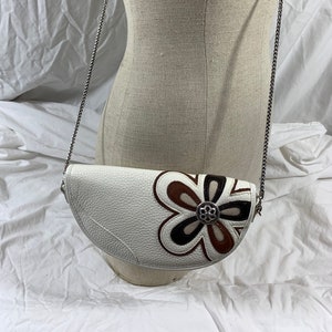 May include: A white leather half-moon shaped crossbody bag with a brown, black, and white flower design. The bag has a silver chain strap.