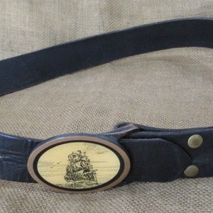May include: A black leather belt with an oval-shaped metal buckle featuring a detailed image of a sailing ship. The buckle is a brass color with a gold-toned background.