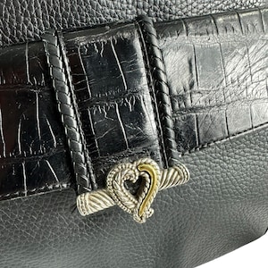 May include: Black leather handbag with a textured finish and a silver and gold heart-shaped clasp. The bag features a braided leather strap detail and a crocodile-embossed flap. The overall design is elegant and classic.