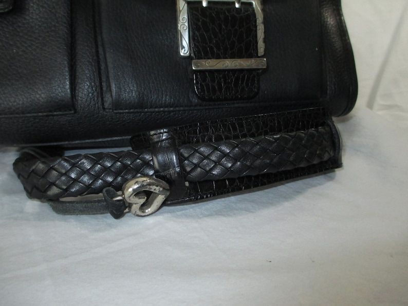 May include: Black leather handbag with a braided strap and a silver heart-shaped charm. The bag has a silver buckle with an intricate design.