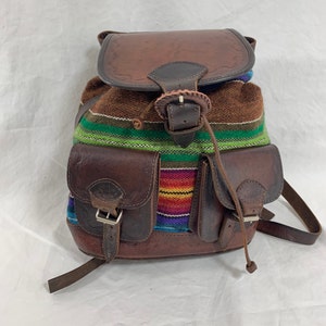 Puede incluir: Una mochila de cuero marrón con dos bolsillos delanteros y cierre de solapa. El cuerpo de la mochila es de tela tejida con rayas coloridas. El cuero tiene detalles en relieve y hebillas metálicas.