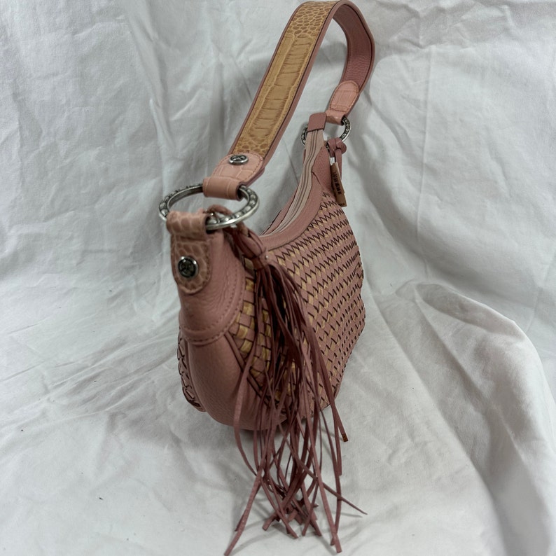 May include: A pink leather shoulder bag with a woven design and fringe detail. The bag features a light brown patterned strap and silver-tone hardware. The bag's dimensions are approximately 12 inches wide and 8 inches tall.