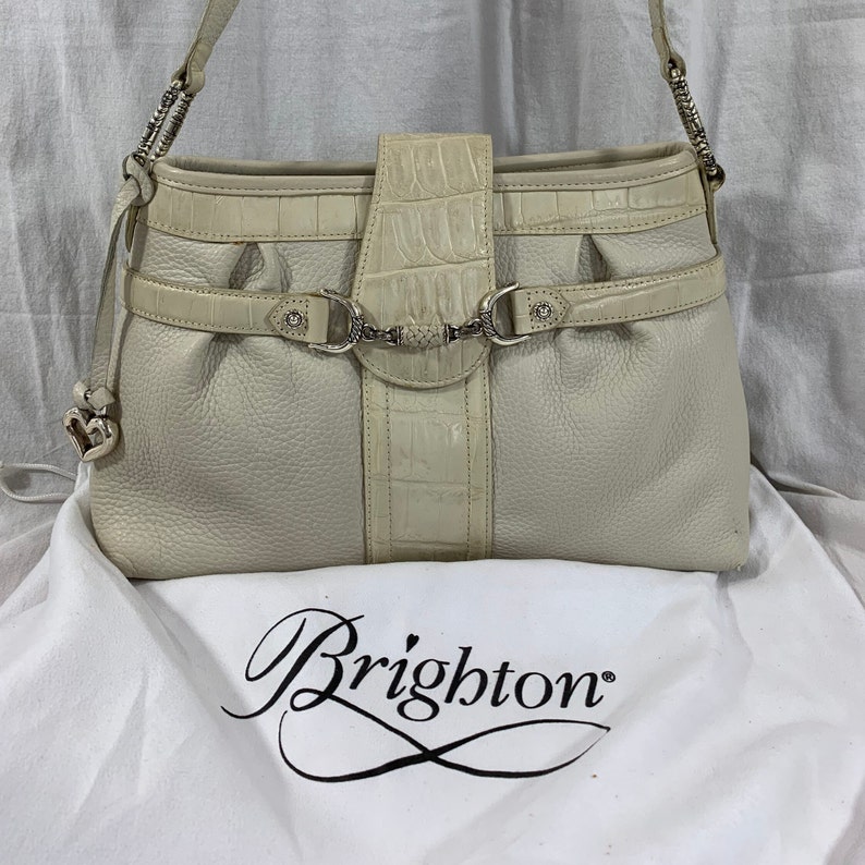 May include: A white leather Brighton handbag with a crocodile embossed design and silver hardware. The bag has a top handle and a shoulder strap.