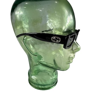 May include: Black sunglasses with a rectangular frame and the Gucci logo on the temples. The sunglasses are displayed on a green glass head. The glasses have a sleek, modern design.