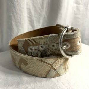 May include: A white leather belt with a silver buckle and a geometric pattern. The belt is made of different pieces of leather sewn together.