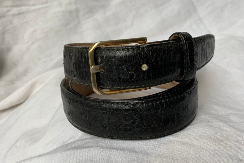Op de afbeelding: Een zwarte leren riem met een gouden gesp. De riem heeft een getextureerd, gegraveerd patroon.