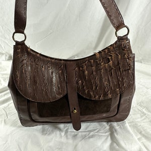 Könnte beinhalten: Eine braune Lederschultertasche mit Klappenverschluss und einem strukturierten Design. Die Tasche hat einen langen verstellbaren Riemen und eine kleine Tasche auf der Vorderseite.