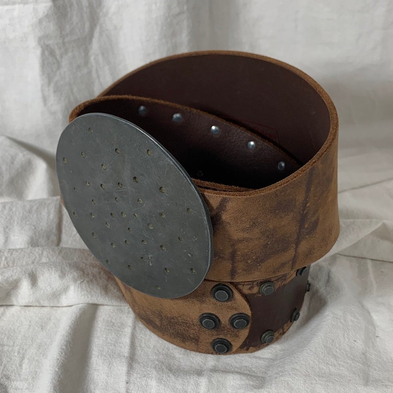 May include: A brown leather belt with a metal buckle and rivets. The buckle has a circular design with small holes.