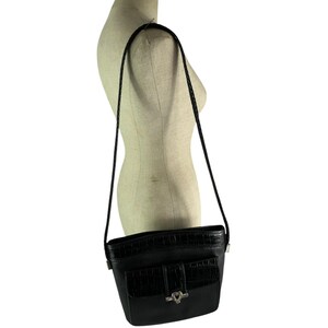 May include: Black crossbody bag with a textured front pocket and a silver-tone heart-shaped clasp. The bag has a braided strap and is displayed on a mannequin. The bag is approximately 8 inches wide and 7 inches tall.