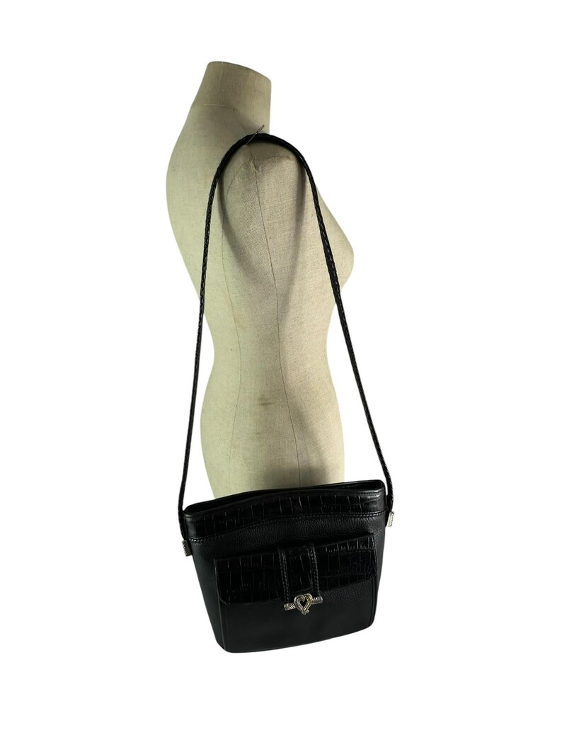 May include: Black crossbody bag with a textured front pocket and a silver-tone heart-shaped clasp. The bag has a braided strap and is displayed on a mannequin. The bag is approximately 8 inches wide and 7 inches tall.