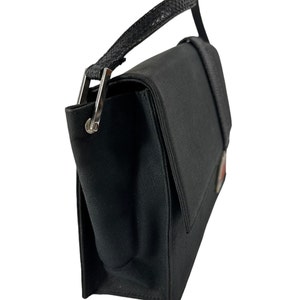 May include: Black handbag with a flap closure and a top handle. The bag features a textured handle and silver-tone hardware. A small emblem is visible on the front. The bag is made of a sturdy material.
