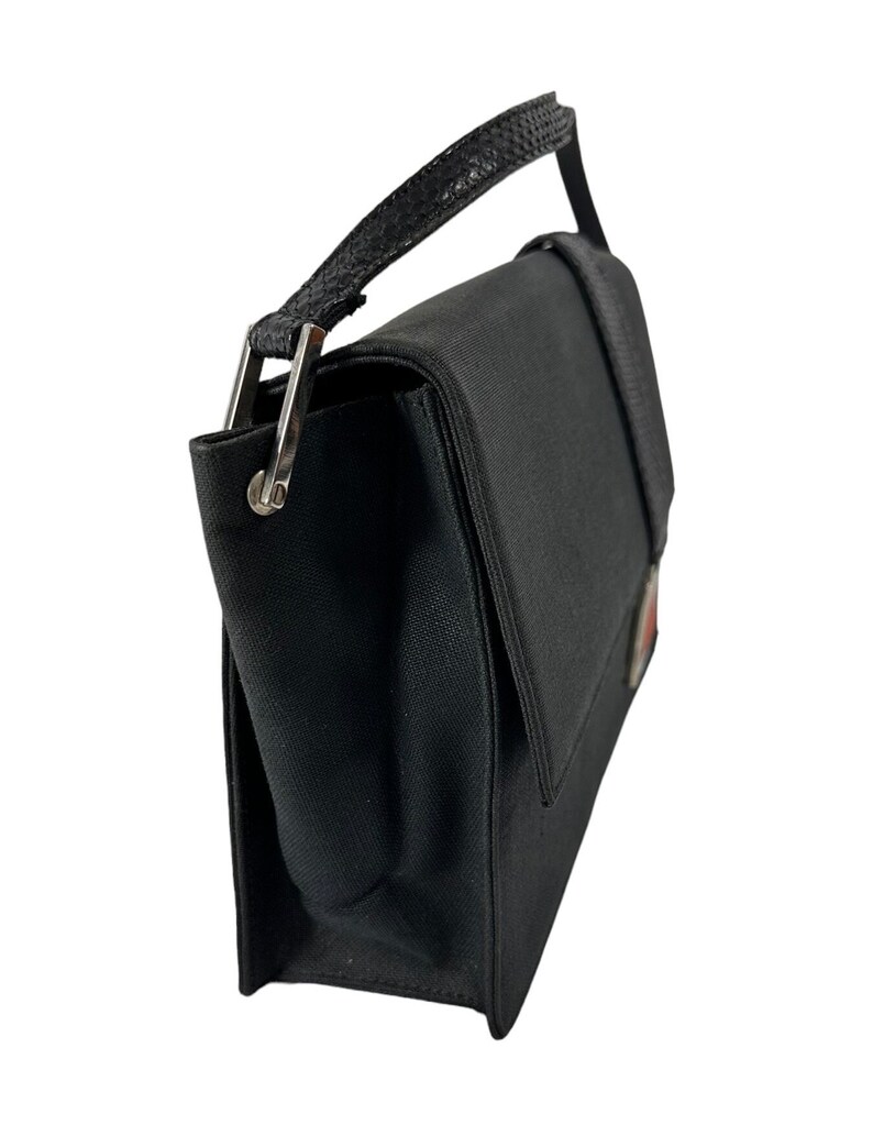 May include: Black handbag with a flap closure and a top handle. The bag features a textured handle and silver-tone hardware. A small emblem is visible on the front. The bag is made of a sturdy material.