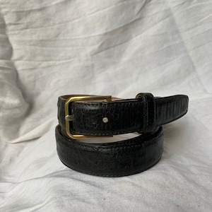 Op de afbeelding: Een zwarte leren riem met een gouden gesp. De riem is dubbelgevouwen en de gesp is zichtbaar.