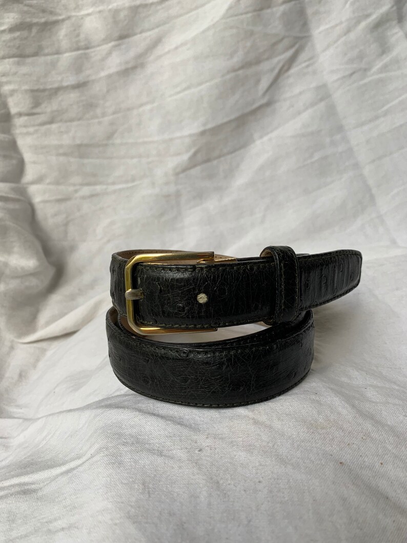 Op de afbeelding: Een zwarte leren riem met een gouden gesp. De riem is dubbelgevouwen en de gesp is zichtbaar.