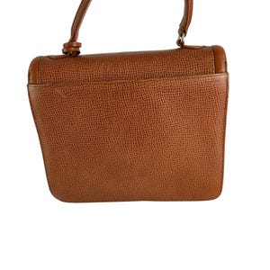Op de afbeelding: Een bruine leren handtas met een textuur. De tas heeft een klepsluiting en een handvat aan de bovenkant. De hardware is goudkleurig. De tas is van een gemiddelde grootte.