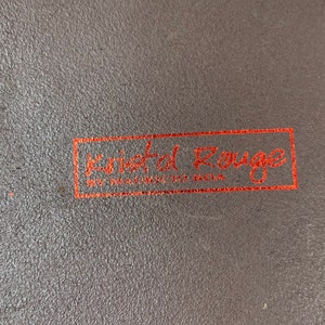 May include: Close-up of a brown leather surface with a red rectangular label that reads "Kristal Rouge" with the words "Handmade in Berlin" underneath.