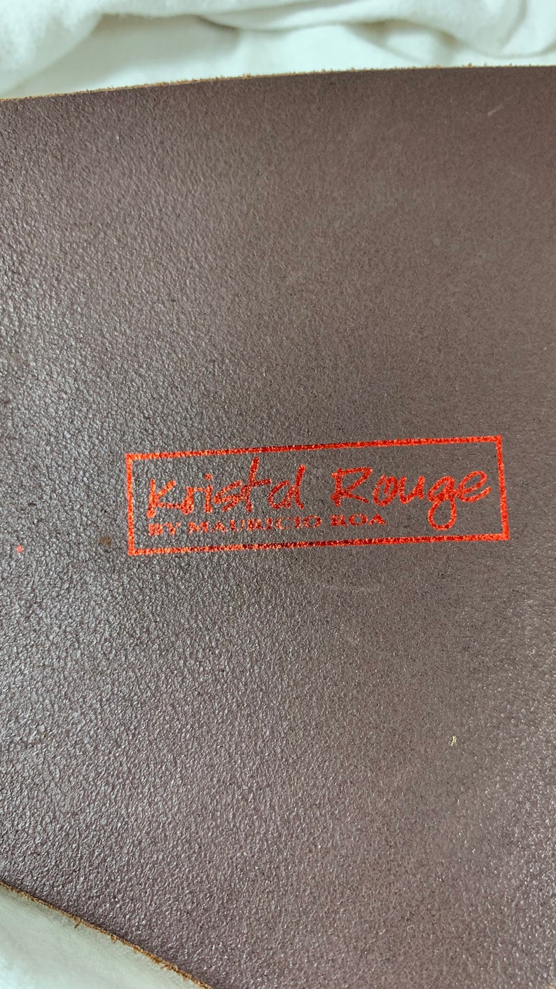 May include: Close-up of a brown leather surface with a red rectangular label that reads "Kristal Rouge" with the words "Handmade in Berlin" underneath.