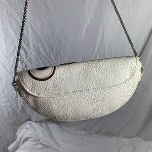 May include: White leather crescent-shaped handbag with a silver chain strap. The bag has a textured surface and a subtle design detail on the front.