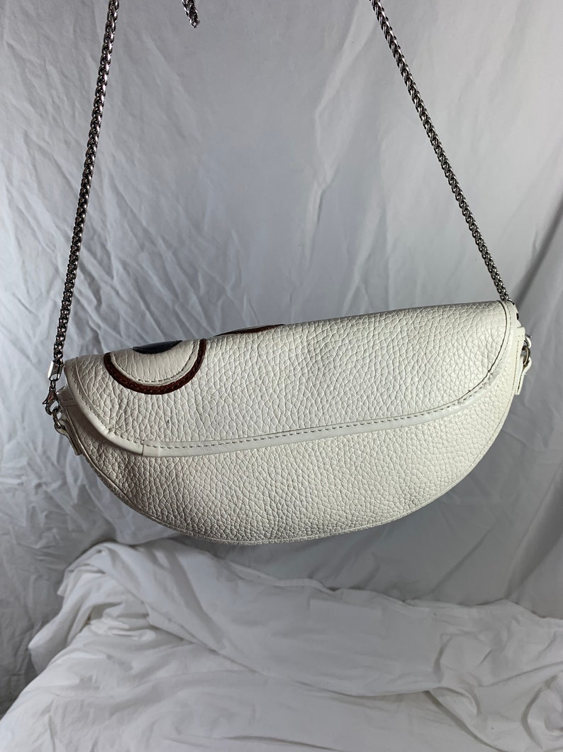 May include: White leather crescent-shaped handbag with a silver chain strap. The bag has a textured surface and a subtle design detail on the front.