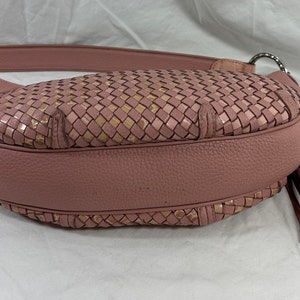 May include: A dusty rose-colored handbag with a woven design on the front. The bag features a leather strap, silver-tone hardware, and a tassel. The woven pattern incorporates a mix of pink and tan tones.