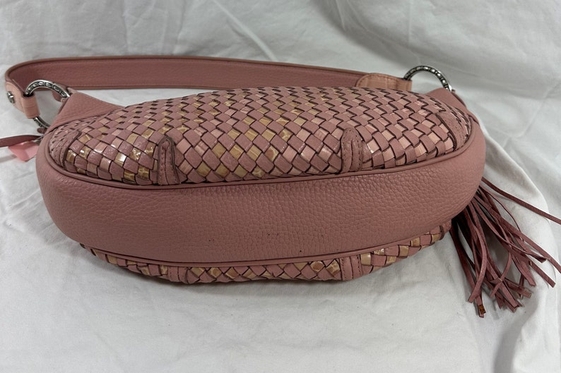 May include: A dusty rose-colored handbag with a woven design on the front. The bag features a leather strap, silver-tone hardware, and a tassel. The woven pattern incorporates a mix of pink and tan tones.
