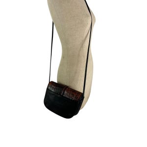 Op de afbeelding: Zwarte crossbodytas met een bruine, getextureerde flap en riem. De tas hangt aan een zwarte riem. De tas wordt op een mannequin gepresenteerd.