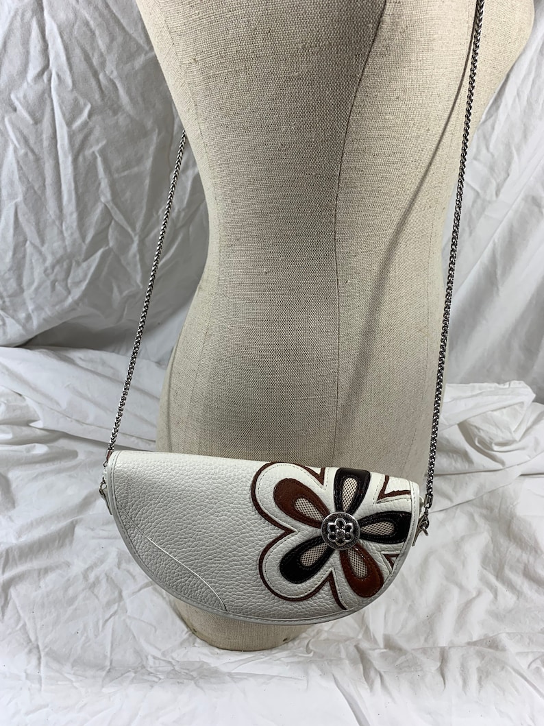 May include: A white leather half-moon shaped crossbody bag with a brown, black, and white flower design. The bag has a silver chain strap.