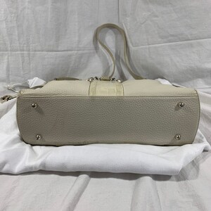 May include: A cream-colored leather tote bag with a crocodile embossed detail on the front. The bag has a top zipper closure and a single strap. The bag is sitting on a white surface.